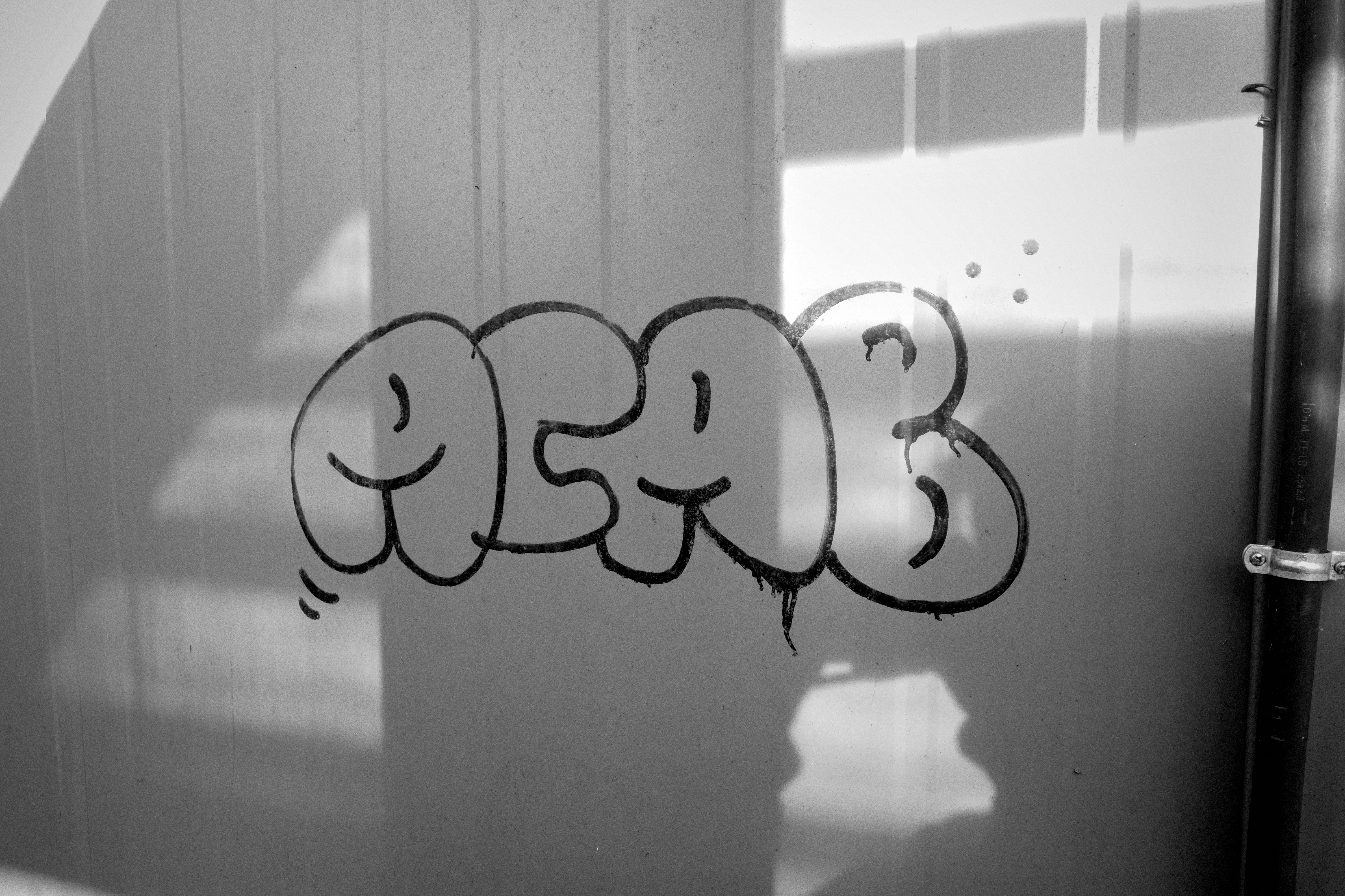 Schwarzweißfoto einer hellen, leicht geriffelten Metallfläche. In der Mitte steht in dunklem, rundem Bubble-Graffiti deutlich der Schriftzug „ACAB“. Die Buchstaben sind weich und zusammenhängend gezeichnet; einige Formen wirken wie einfache Gesichter oder Smileys, besonders links und im mittleren Bereich. Rechts am Schriftzug sind kleine Punkte sowie verlaufende Farbtropfen zu sehen. Starke Schatten fallen diagonal und rechteckig über die Wand, vermutlich von einem Fenster, Geländer oder einer Konstruktion. Am rechten Bildrand verläuft ein dunkles senkrechtes Rohr mit einer hellen Metallschelle. Das Bild wirkt urban, grafisch und kontrastreich.