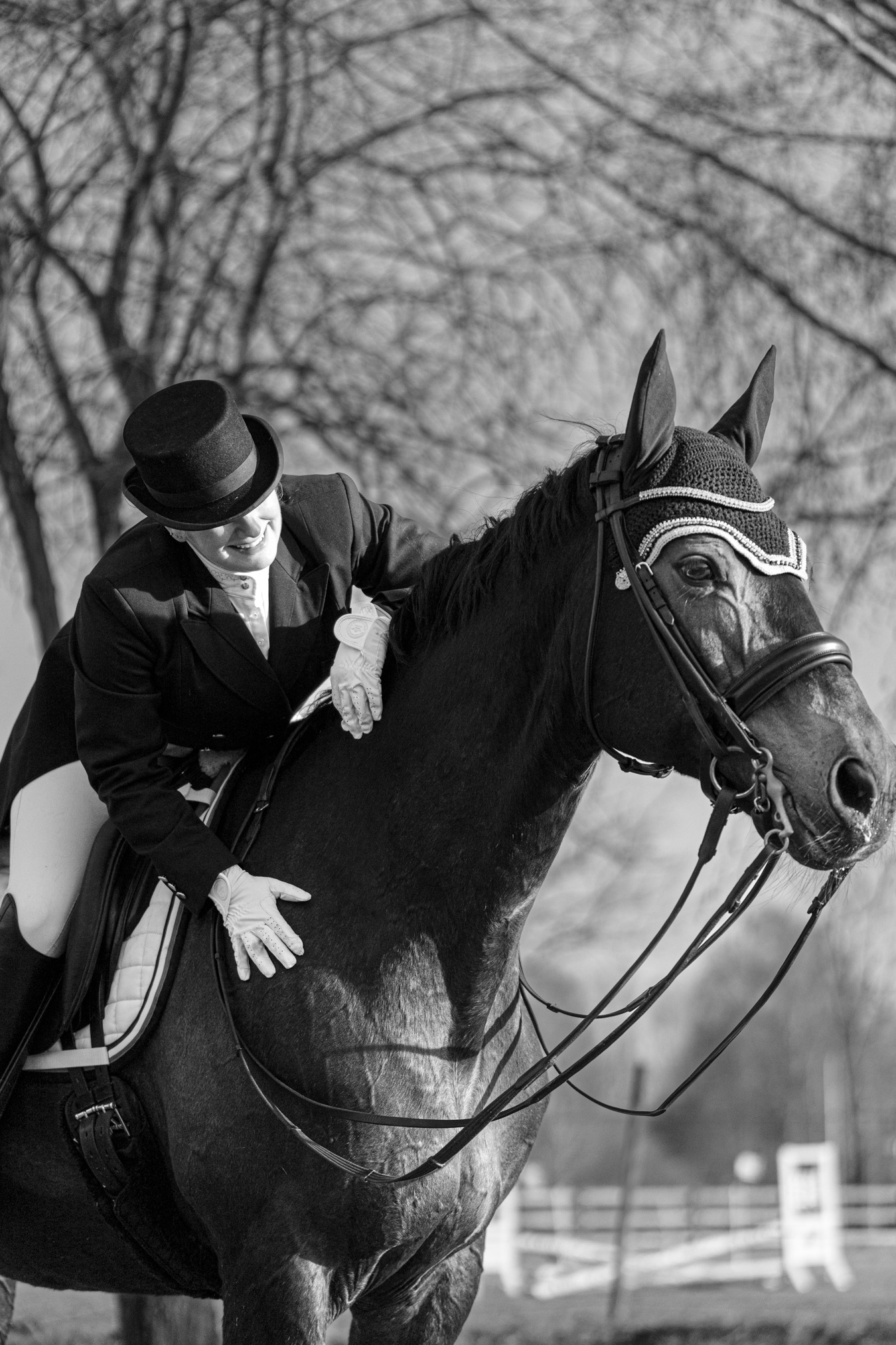 Eine Reiterin in eleganter, schwarzer Reitbekleidung mit Zylinder sitzt auf einem majestätischen, dunklen Pferd. Ihre weiße Reithose und die weißen Handschuhe kontrastieren stark mit dem dunklen Anzug. Sie beugt sich vor und streichelt das Pferd liebevoll am Hals, was eine innige Verbindung zwischen Mensch und Tier vermittelt. Das Pferd trägt eine verzierte Trense und eine schützende Ohrenhaube. Im Hintergrund sind kahle Bäume zu erkennen, deren Äste filigrane Muster gegen den Himmel bilden. Die schwarz-weiße Farbgebung des Bildes verleiht der Szene eine zeitlose Eleganz und hebt die Details der Kleidung und des Pferdes hervor. Die Stimmung wirkt ruhig und harmonisch, während das Licht sanft auf die Szene fällt und die Konturen betont. [KI-generiert]